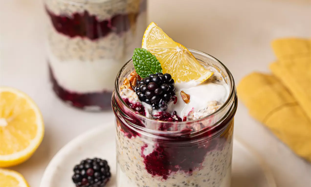 Picture - Layered lemon and chia seed oats in glass jars with swirls of berry compote, cream topping, blackberries, lemon slices, and mint leaves