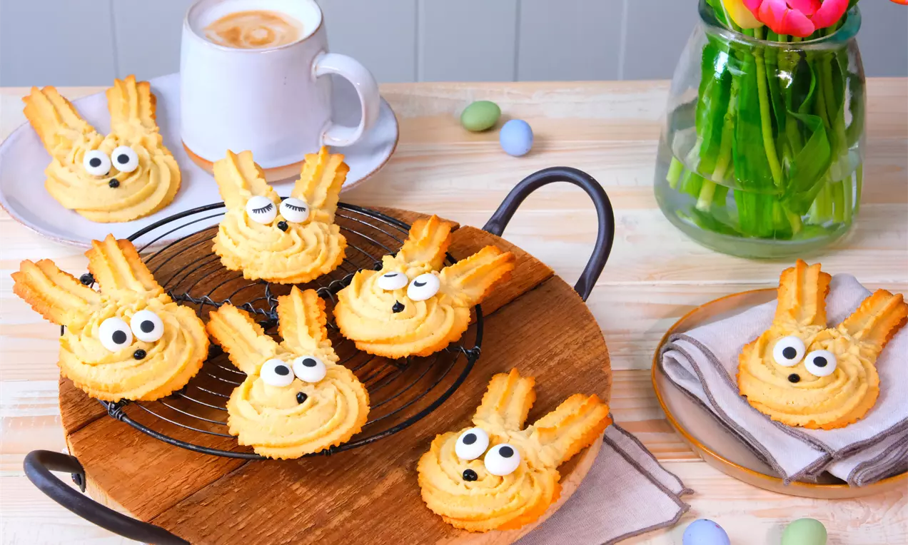 Biscotti dorati a forma di coniglietto con occhi di zucchero spruzzati su una griglia nera, su un tagliere di legno circondati da tulipani e uova colorate
