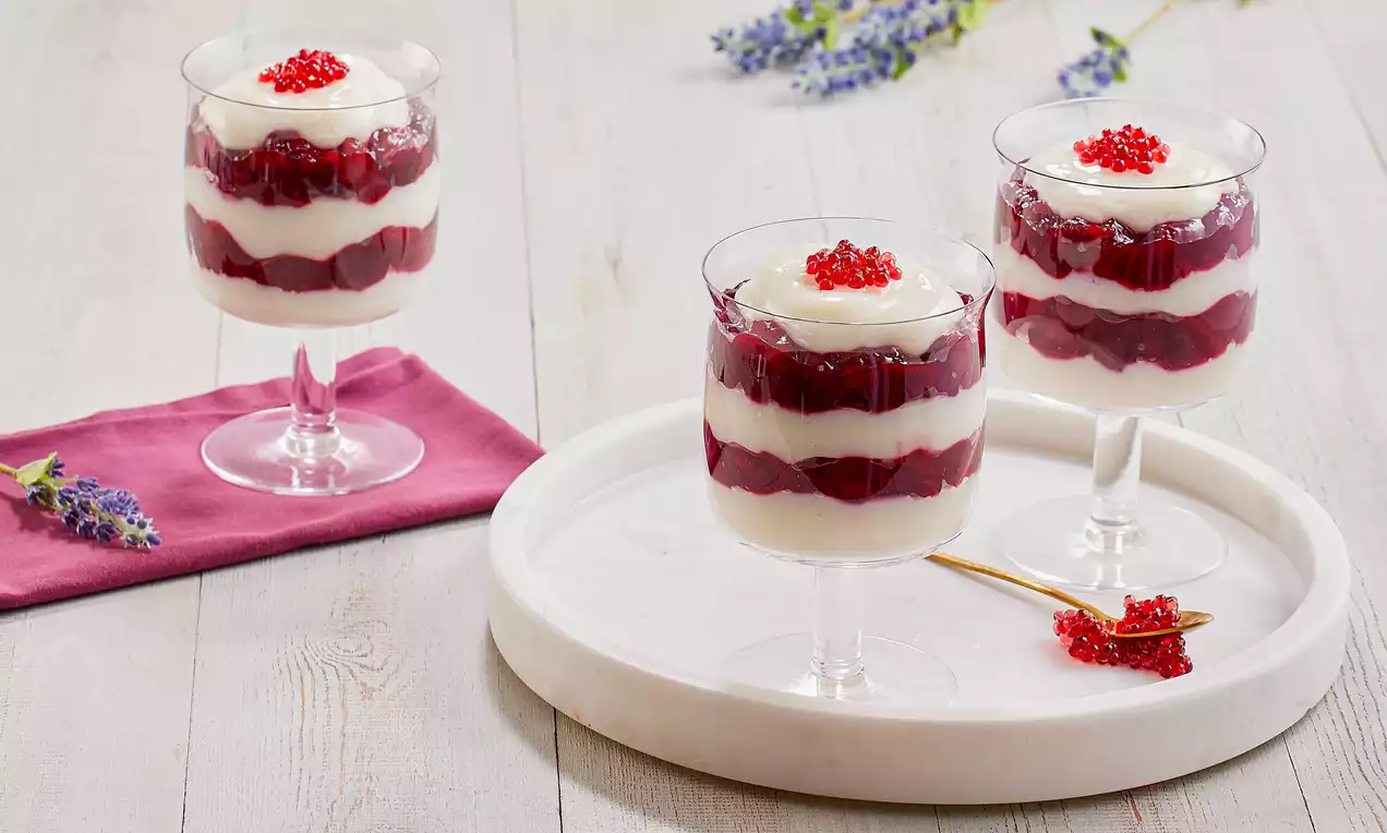 Cam ayaklı kupalarda, kat kat dizilmiş beyaz puding ve parlak kırmızı vişnelerle, üstü kırmızı süslemeli zarif bir sunum