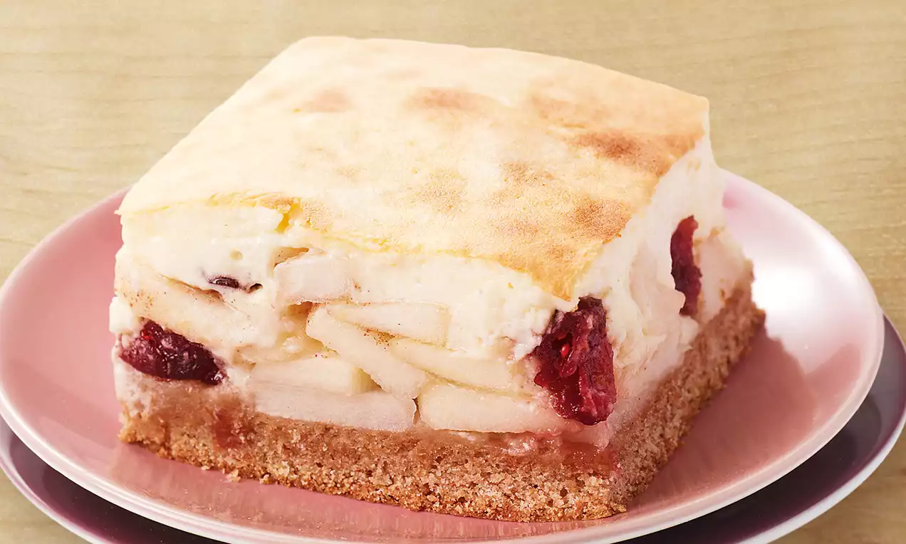 Carré de gâteau avec une base biscuitée brune, couches de pomme pâles et morceaux rouges nichés sous une crème légère sur assiette rose
