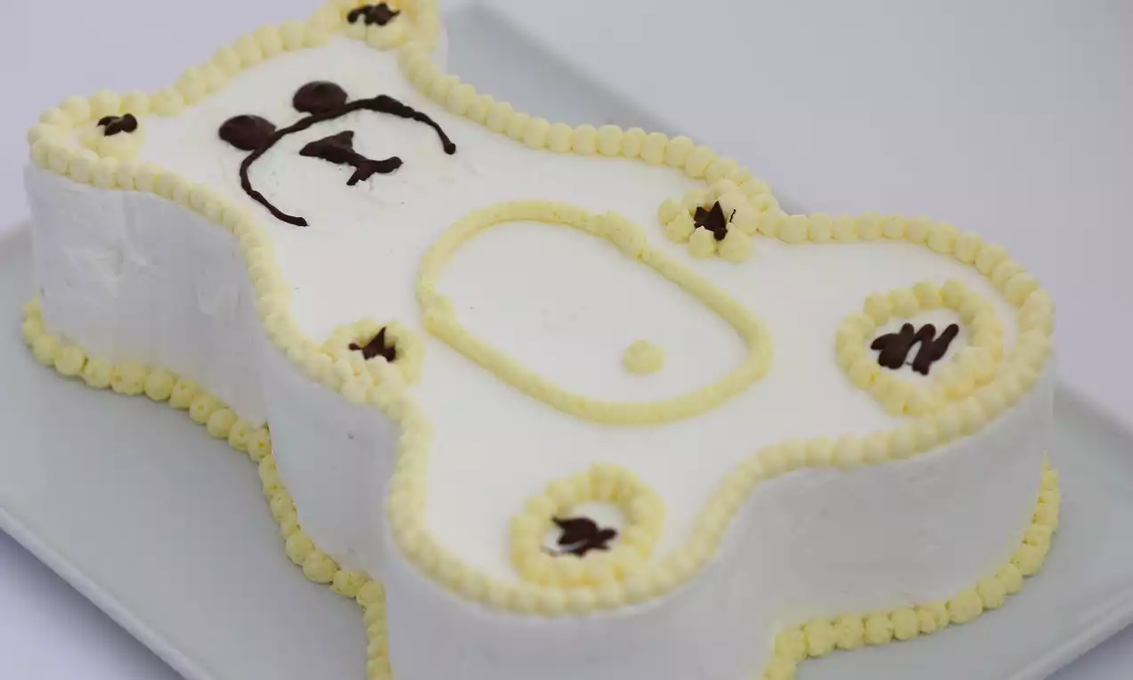 Beyaz kremayla kaplı ayıcık şeklindeki pasta, sarı şanti şeritleri ve çikolatalı detaylarla süslenerek dikdörtgen bir tabakta sunulmuş