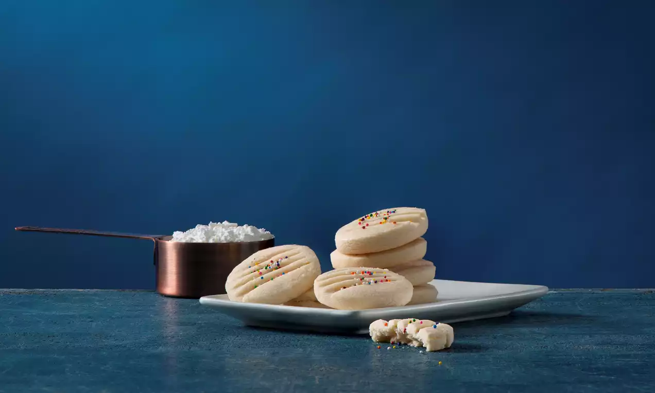 Soft, pale yellow melting moments with delicate ridges, sprinkled with colorful nonpareils on a white plate against a deep blue backdrop