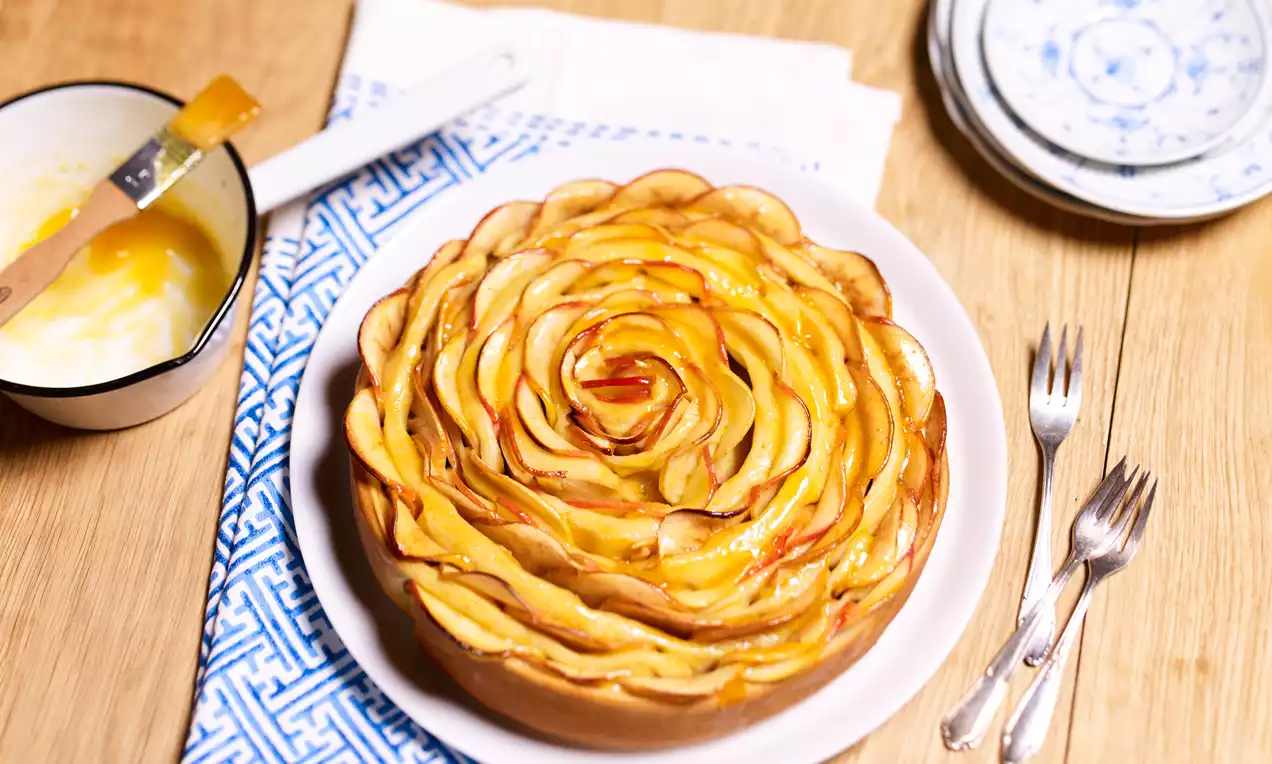 Gâteau aux pommes dorées disposées en fines lamelles formant une rose, sur une assiette blanche avec des fourchettes argentées et une nappe bleue