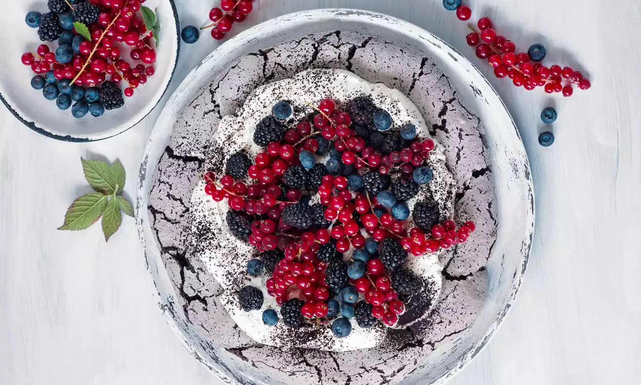 Pavlova s popraskaným černým kakaovým základem, nadýchanou bílou pěnou zdobenou lesklými červenými ríbezlemi, ostružinami a borůvkami