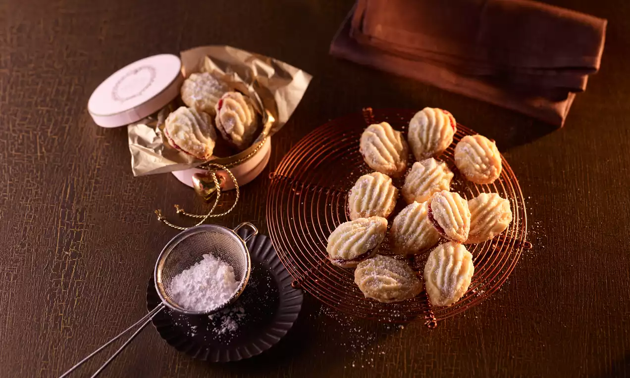 Goldgelbes Spritzgebäck mit geriffelter Oberfläche, teils mit Puderzucker bestäubt, auf rotem Kuchengitter und in rundem Geschenkkarton