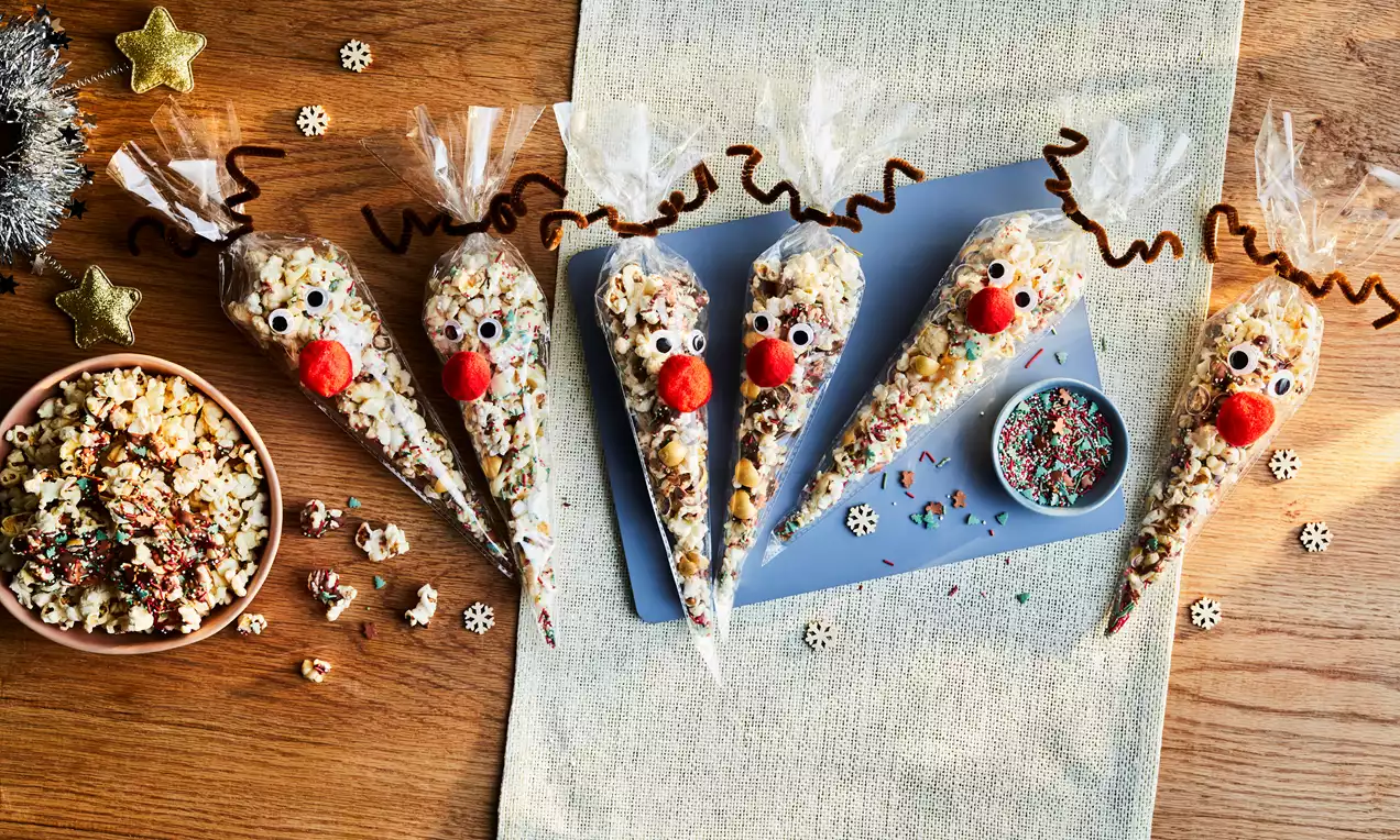 Clear cone-shaped bags filled with white and caramel popcorn, decorated as reindeer with red noses, googly eyes and brown antlers on a wooden table with a beige cloth, sprinkles and scattered decorative stars