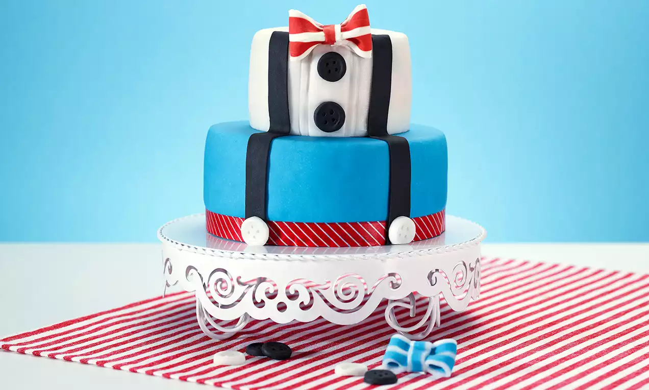 Zweistöckige Torte mit blauem Fondant, schwarzen Hosenträgern, roten und weißen Streifen, Knöpfen und rot-weißer Schleife auf weißer Tortenplatte