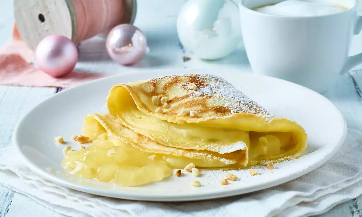 Goudgele gevulde pannenkoek met vulling die uitloopt, bestrooid met poedersuiker en nootjes op een wit bord, met winterdecoratie op de achtergrond