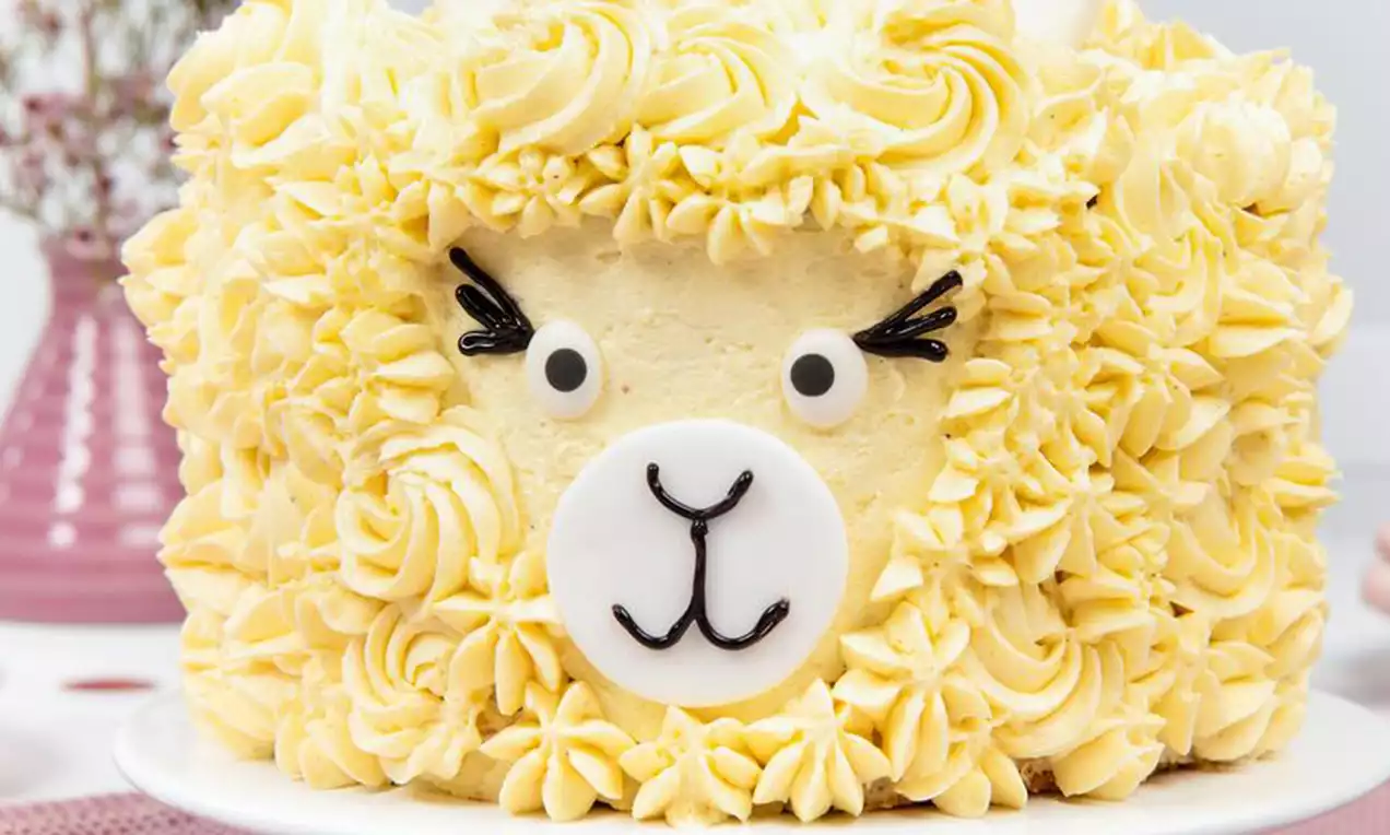 Eine gelbe Torte in Form eines Lamas mit flauschiger Creme-Textur, Gesicht aus Fondant und Zuckerdekor auf weißer Tortenplatte