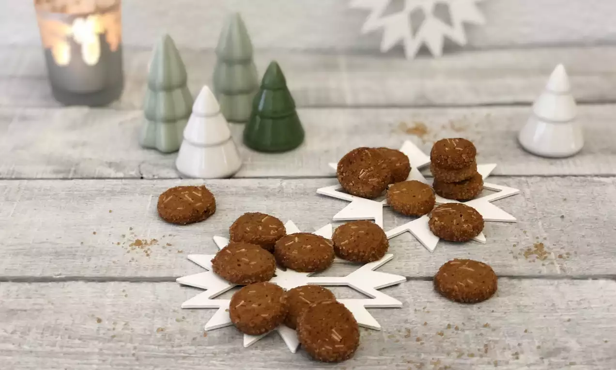 Kleine, goldbraune Gewürzkekse mit körniger Textur auf weißen sternförmigen Untersetzern, umgeben von dekorativen Weihnachtsbäumen