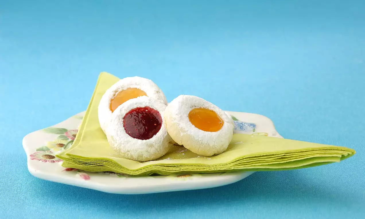 Renkli marmelat dolgulu beyaz pudra şekerli yuvarlak kurabiyeler, desenli porselen tabakta sarı peçete üzerinde düzenlenmiş