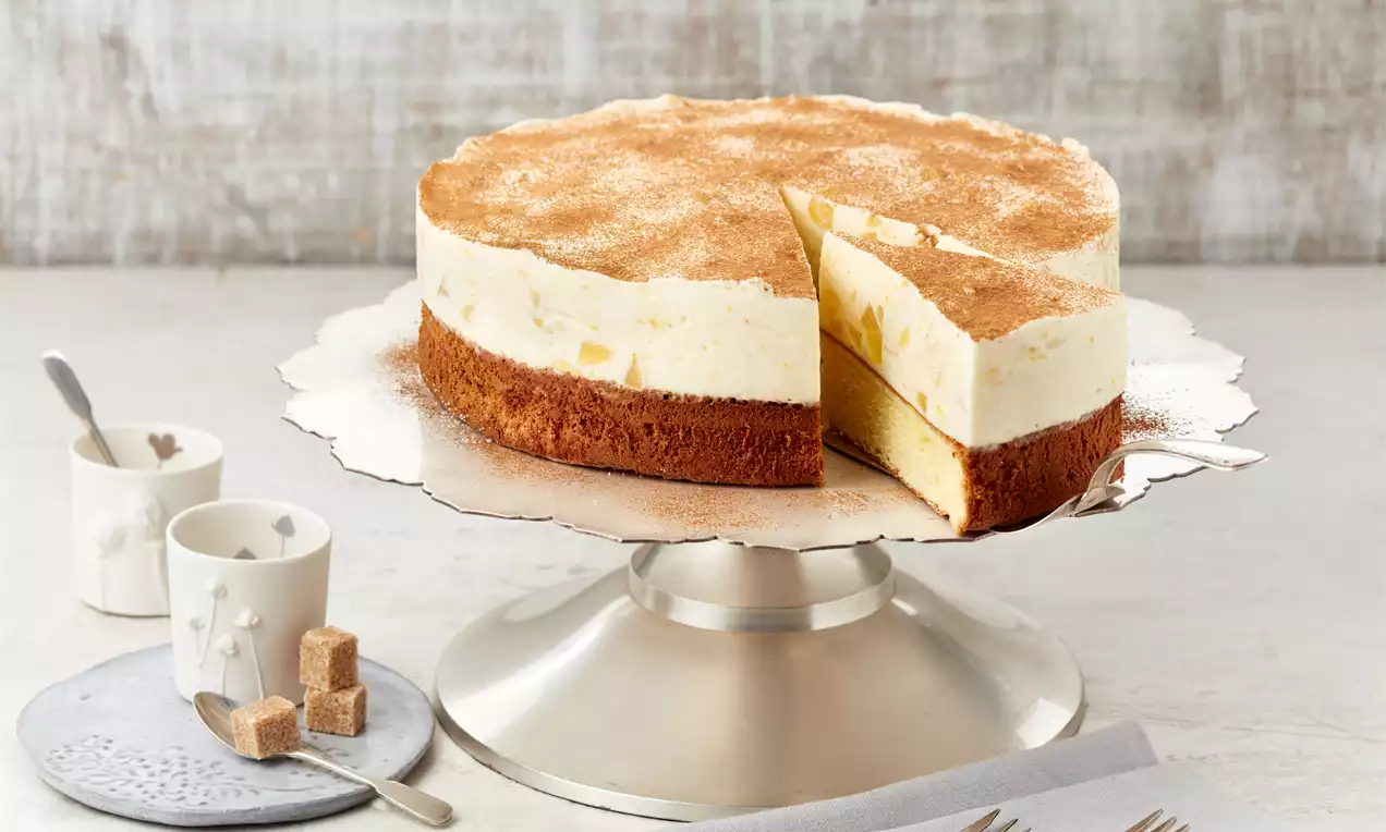 Winterapfel-Torte mit braunem Boden und heller Creme, dünn mit Zimt bestreut, auf silberner Tortenplatte mit Anschnitt und gedecktem Tisch