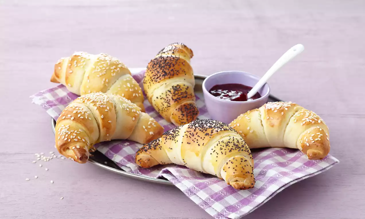 Goldgelb gebackene, filigrane Hörnchen mit Mohn und Sesam bestreut auf kariertem Tuch, dazu eine violette Schale mit roter Marmelade