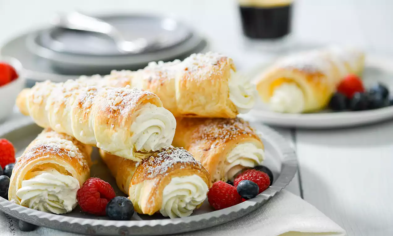 Zlatnosmeđi roščići od lisnatog tijesta punjeni bijelom kremom, posuti šećerom u prahu, servirani na tanjiru s bobičastim voćem