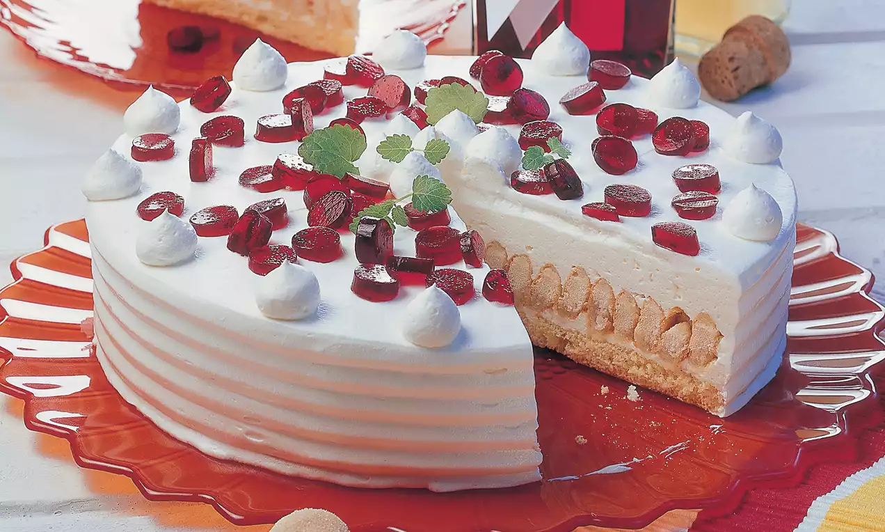 Weiße Holunder-Torte mit glatter Creme, gekrönt von roten Geleestückchen, Sahnetupfen und Minzblättern auf rotem Teller angerichtet