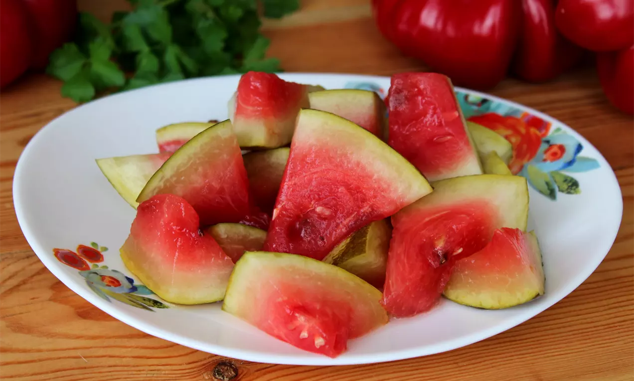 Парчета червено-розова диня с тънка зелена кора, разположени хаотично върху бяла чиния с шарки, на дървен фон с листа магданоз