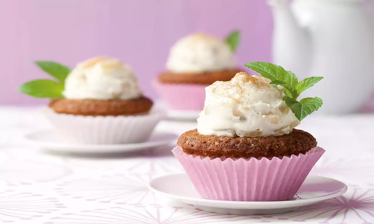 Braune Muffins in rosa Förmchen mit weißer Cremehaube, goldenen Streuseln und grüner Minze auf kleinen weißen Tellern vor lila Hintergrund