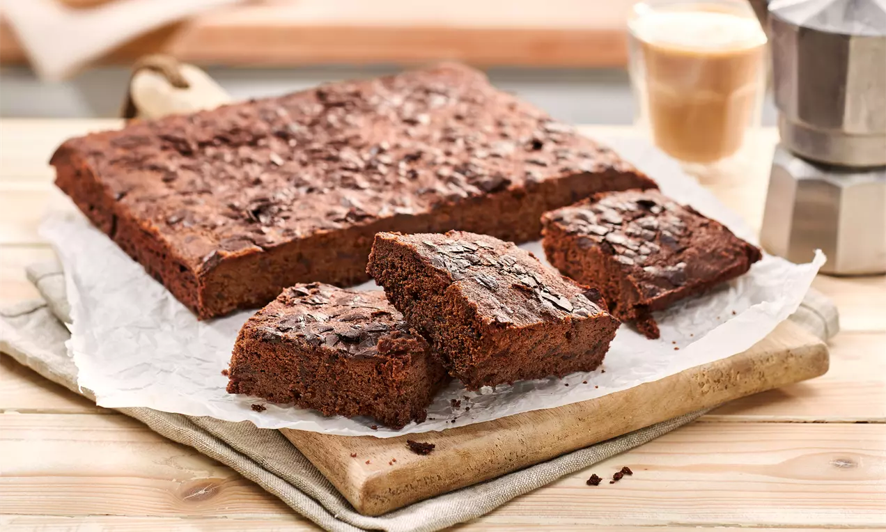 Saftige, dunkelbraune Brownies mit lockerer Textur und glatter Oberfläche auf Backpapier, angerichtet auf einem Holzbrett, im Hintergrund Kaffee und Espressokanne