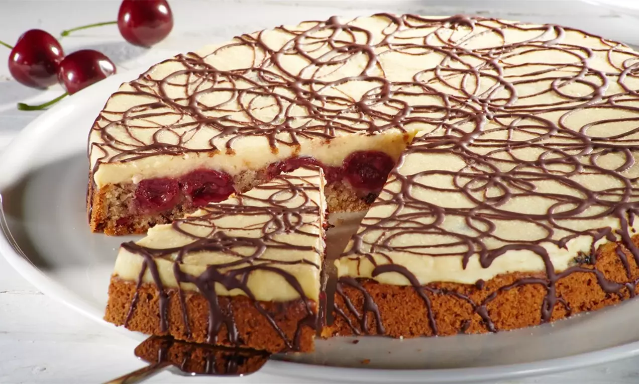 Кръгла марципанова торта с кремообразен светъл слой, покрит с тънки шоколадови нишки, върху кафяв основен блат със сочни череши