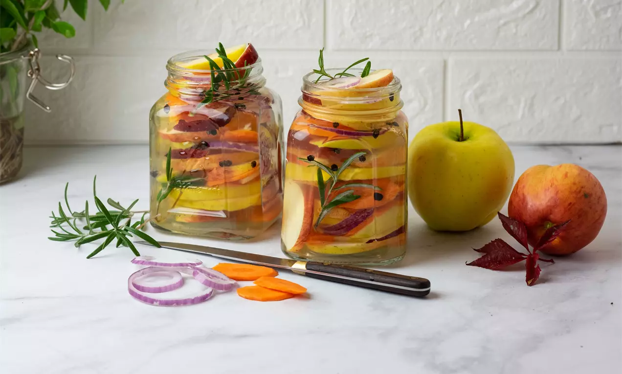 Светложълти и червени ябълки нарязани на тънки резени в стъклени буркани с клонка розмарин, черен пипер и резени морков върху бял плот