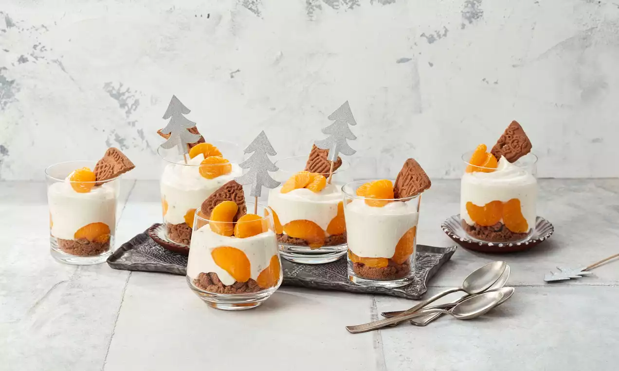 Spekulatius-Dessert in Gläsern mit braunem Keksboden, weißer Creme, orangefarbenen Mandarinenstücken und Dekoration aus Keks und silbernen Tannenbäumen