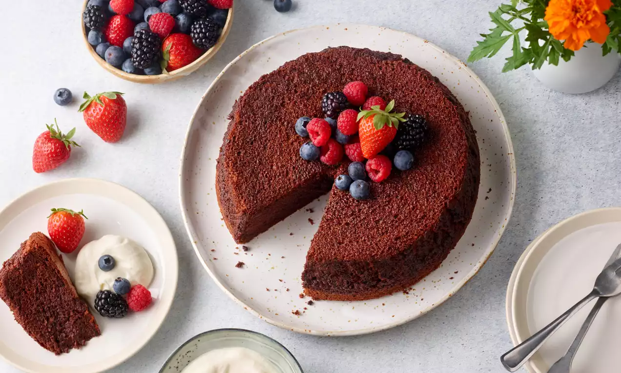 Runder, dunkelbrauner Schokokuchen mit feiner Textur, belegt mit frischen Beeren auf weißem Teller, daneben Sahne und Beeren serviert