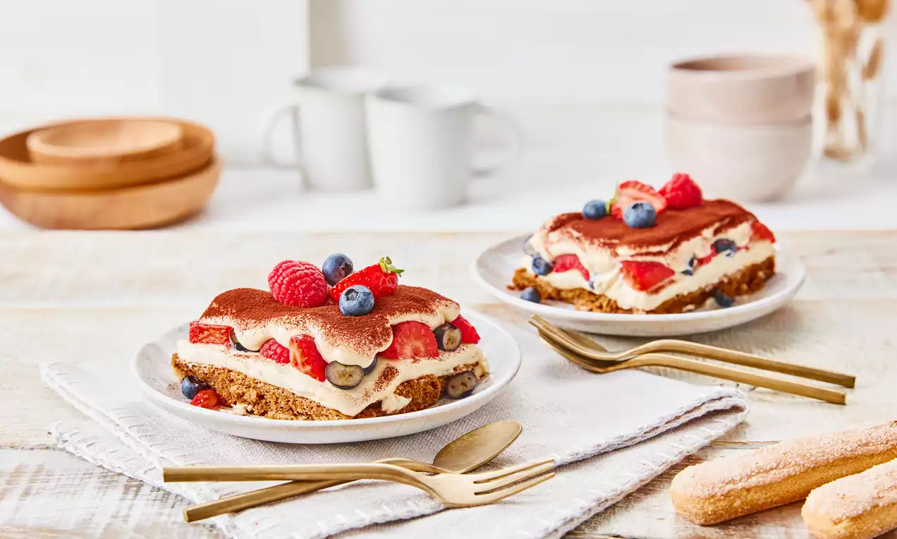 Zwei Portionen Beeren-Tiramisu mit hellen Creme- und Keks-Schichten, roten und blauen Beeren sowie Kakao bestäubt auf weißen Tellern