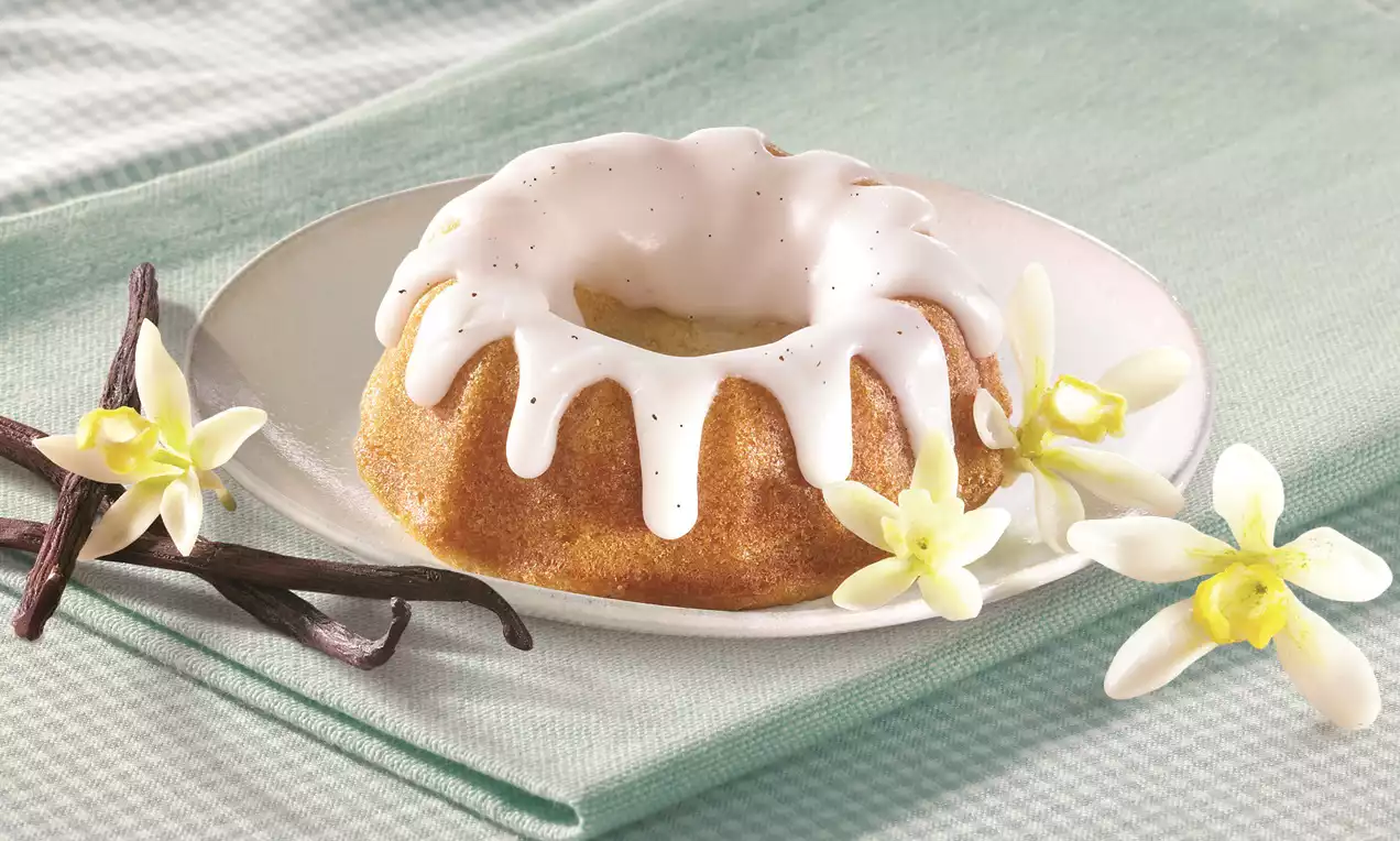 Mini vanilla bundt cake with golden soft texture and glossy white icing, garnished with vanilla pods and delicate white flowers on a white plate