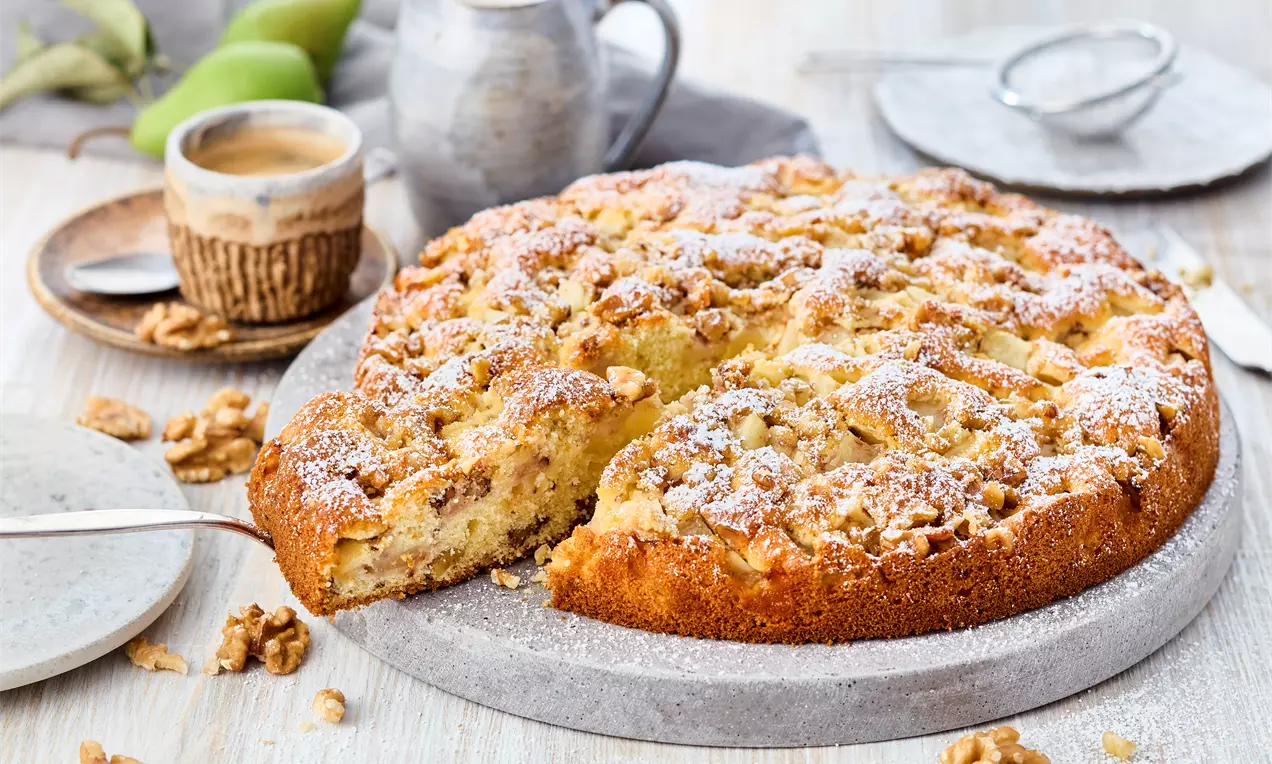 Ein goldbrauner Birnen-Walnuss-Kuchen mit lockerer Kruste, bestäubt mit Puderzucker, serviert auf grauem Steinbrett mit Walnüssen dekoriert