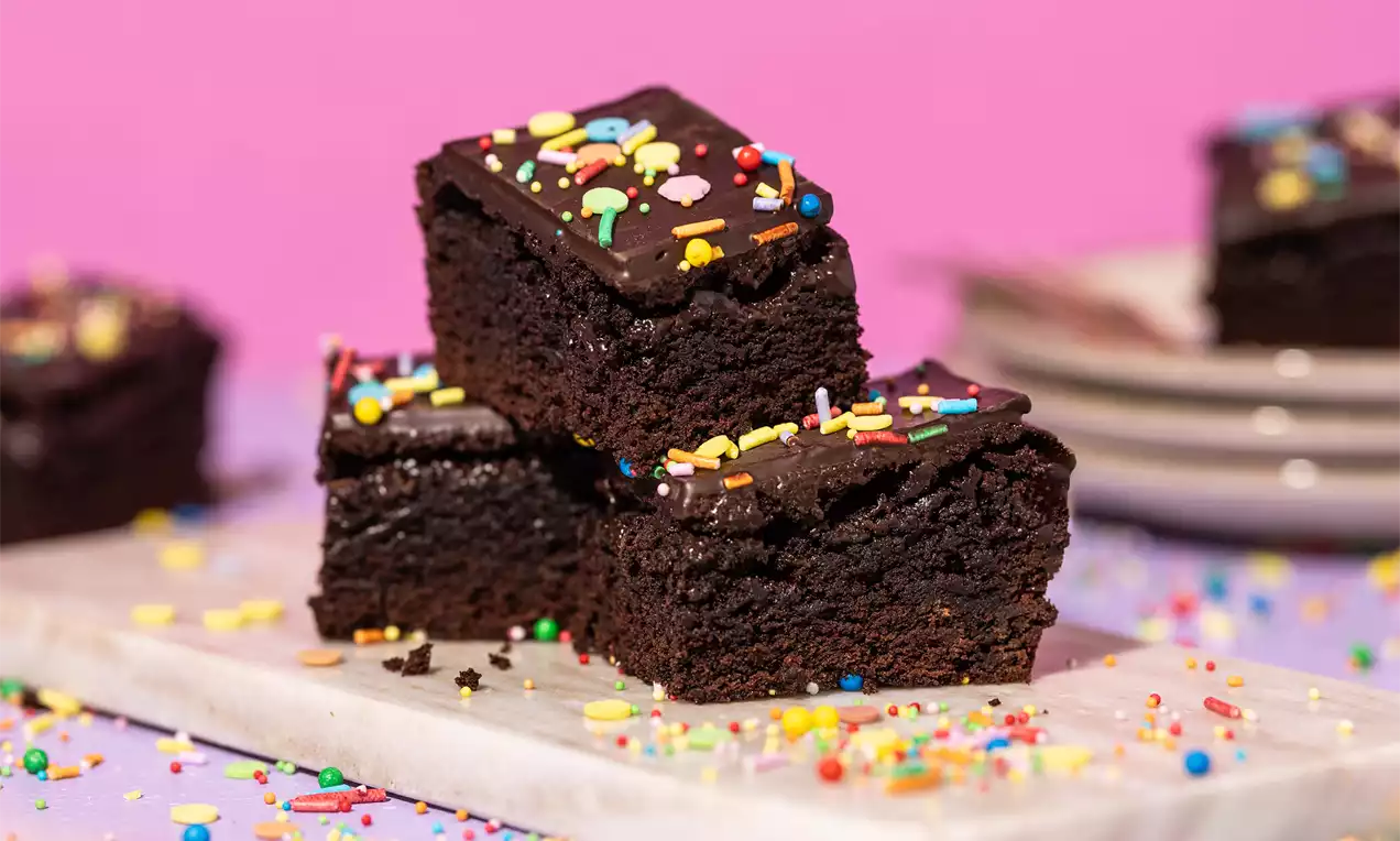 Donkere, vierkante brownies met glanzende chocoladetopping en kleurrijke hagelslag op een marmeren plank tegen een roze achtergrond