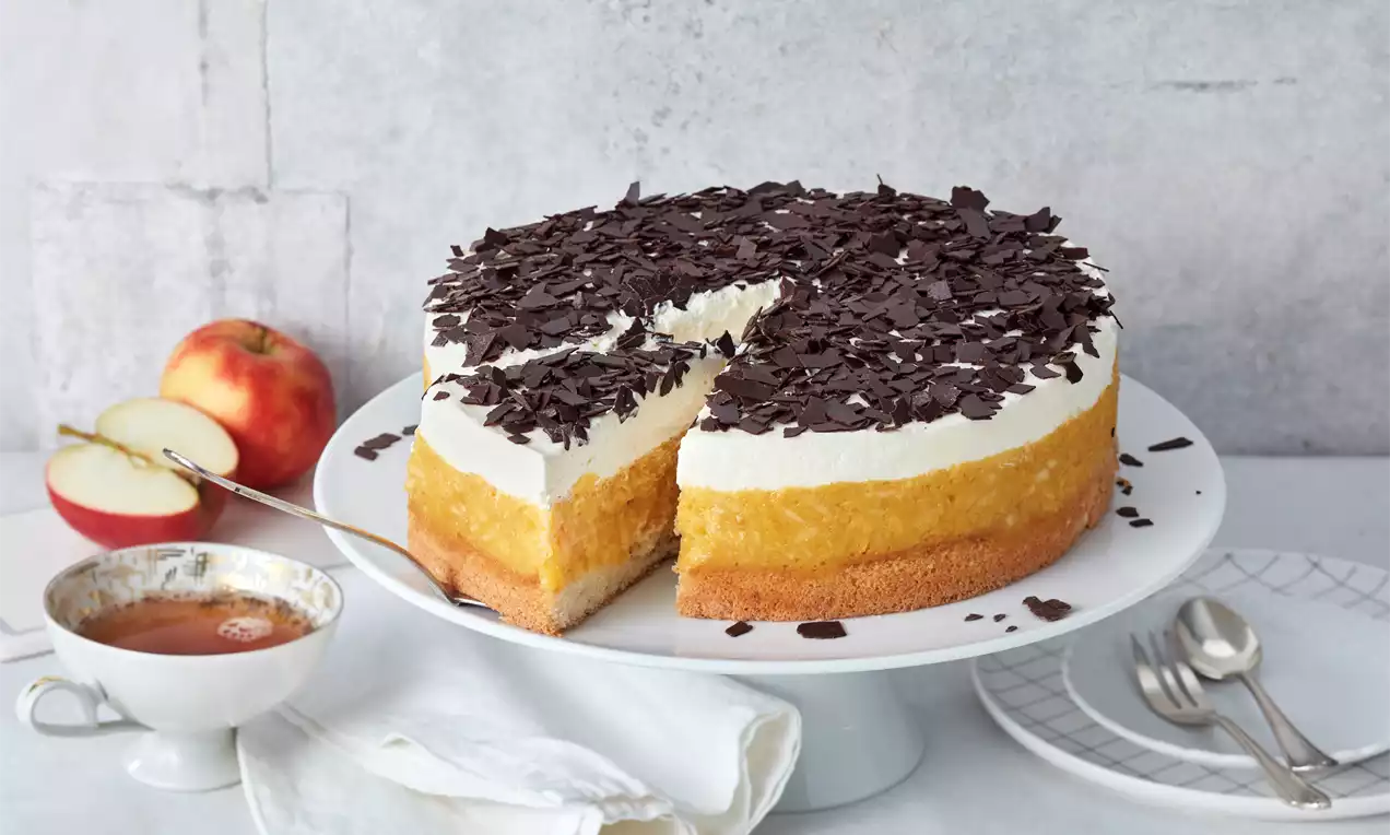 Runde schwedische Apfeltorte mit goldgelber Apfelschicht, weißer Creme und dunklen Schokoraspeln auf weißer Tortenplatte angerichtet