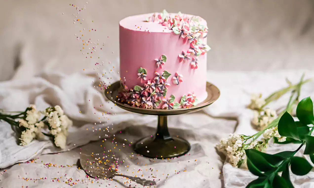 Hortensiatårta med rosa glasyr, dekorerad med små blommor i vitt och rosa, på en mörk tårtställning med gröna blad och vita blommor runtom