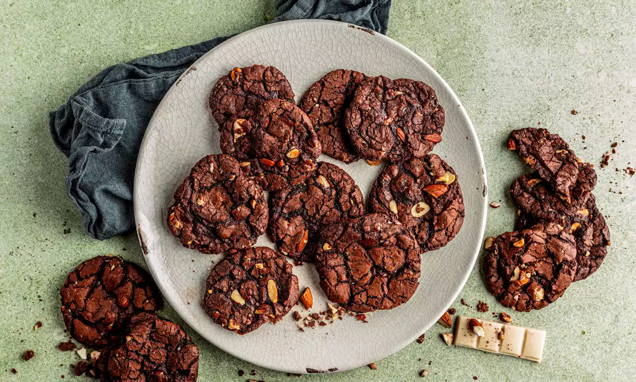 Donkerbruine vegan brownie cookies met een gebarsten textuur en noten, liggend op een lichtgrijs bord met een donkere linnen doek eronder