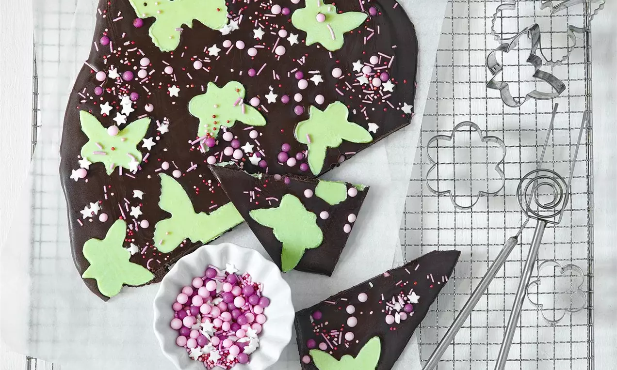 Tmavá nepravidelná placka čokolády zdobená zelenými máslovými motýlky, růžovými a bílými cukrovými kuličkami a posypem na mřížce s formičkami