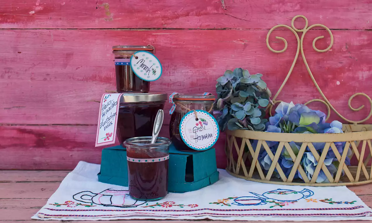 Zwetschgenmus in einem Glas, dunkelviolett glänzend, mit Löffel, frischen Zwetschgenhälften und rustikalem Holzbrett serviert