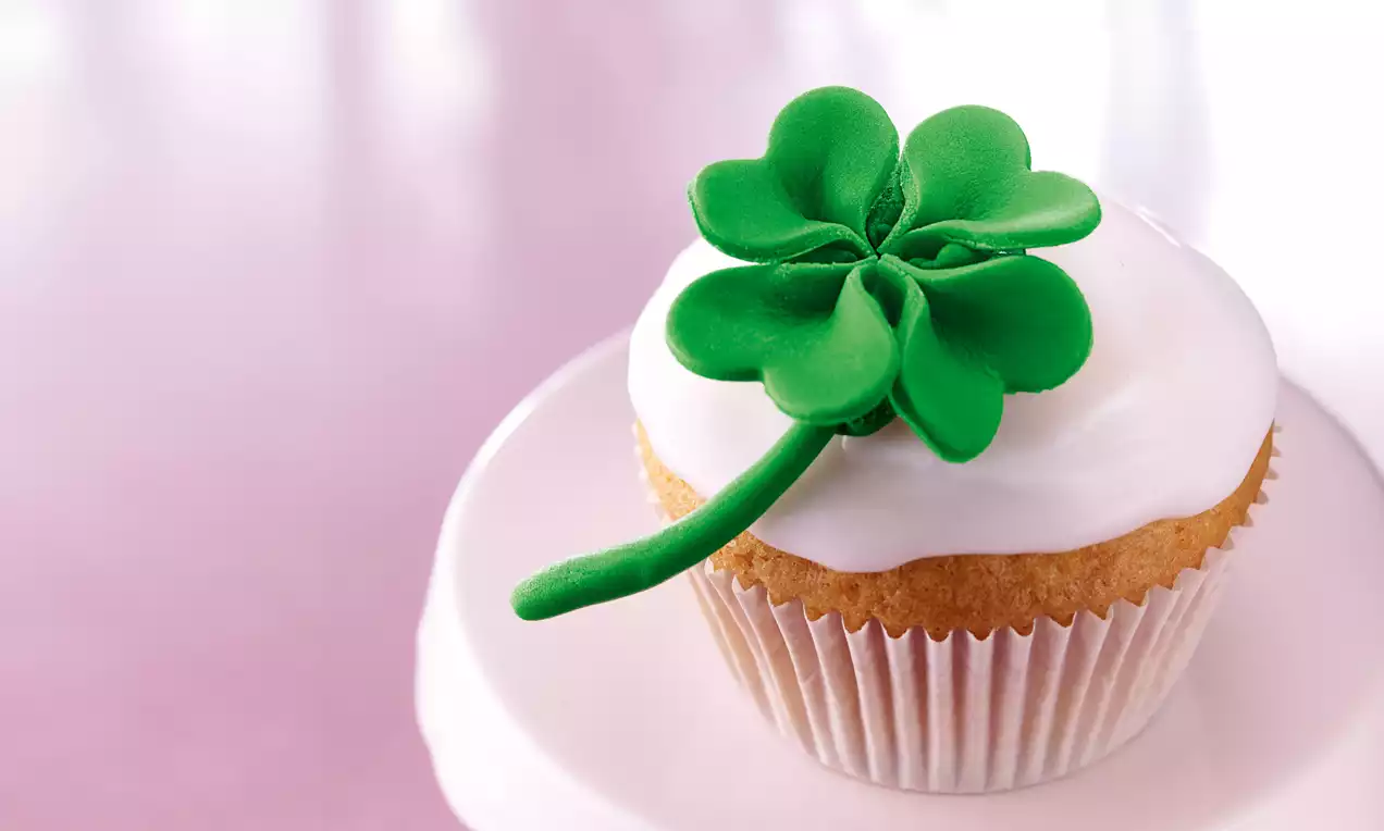 Cupcake med hvit glasur toppet med grønt marsipan-kløverblad, plassert på en lys rosa tallerken mot en myk rosa bakgrunn