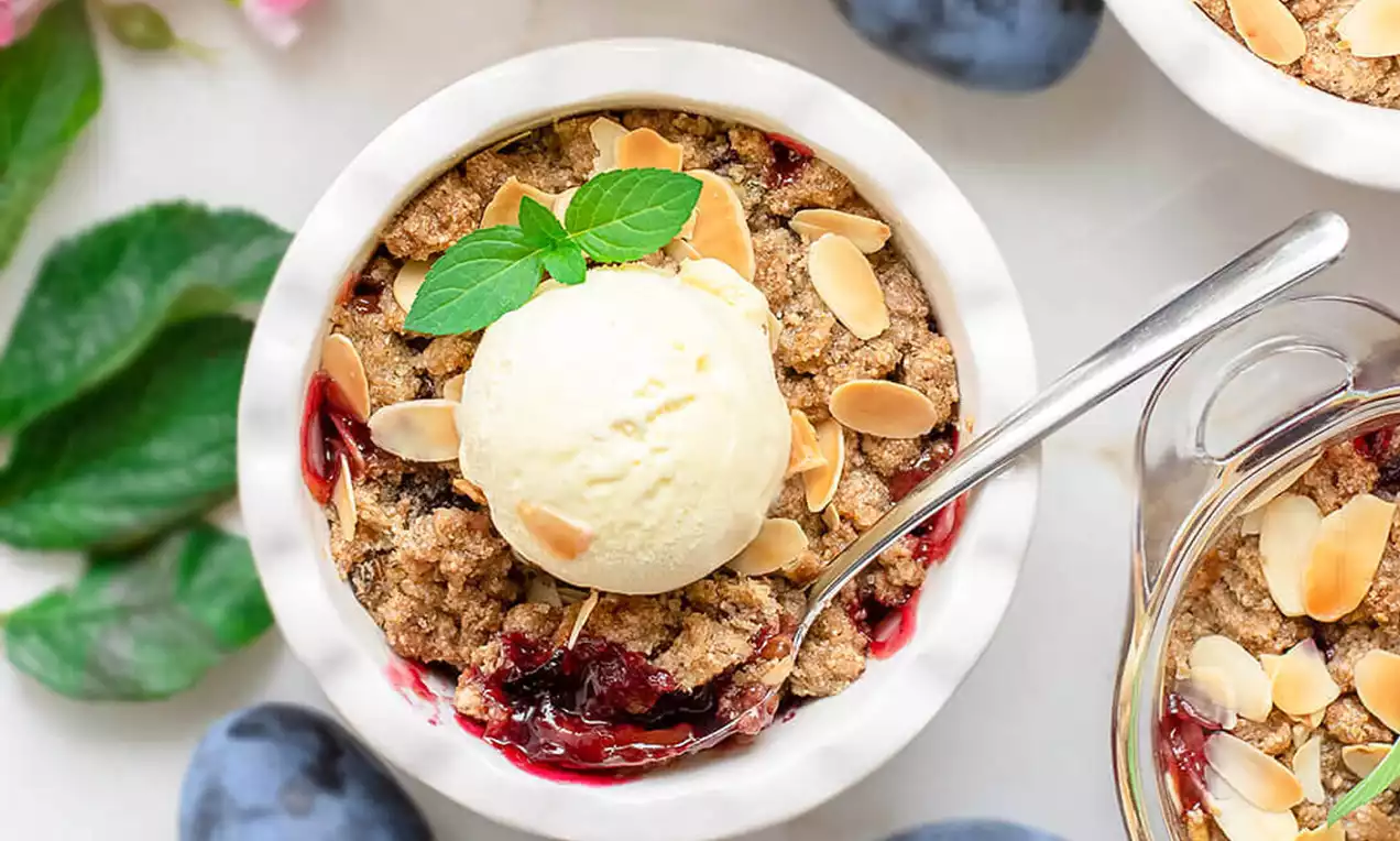 Crumble sa šljivama u bijeloj zdjelici s hrskavom zlatnosmeđom koricom, posut listićima badema i kuglom sladoleda te svježim listićem mente