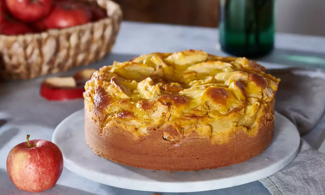 Goldbrauner Apfelkuchen mit welliger Oberfläche auf weißer Marmorplatte, daneben ein roter Apfel und Korb voller Äpfel im Hintergrund