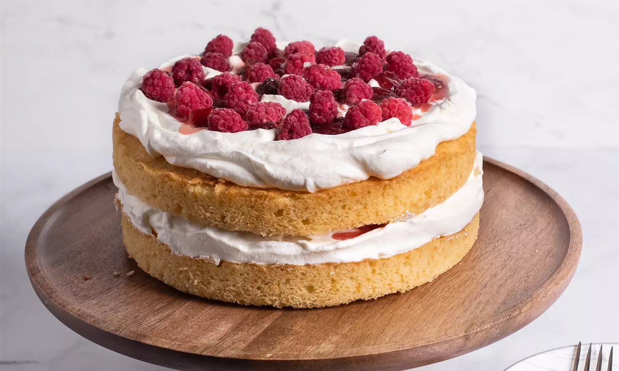 Светли пухкави блатове, слоени със сметана и фин пласт сладко, гарнирани с ярки червени малини, върху дървена поставка