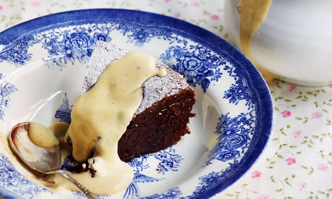 Trekanthakket sjokoladekake med mørkebrun farge og pudret melis, servert med lys gul crème anglaise på et blåblomstret fat