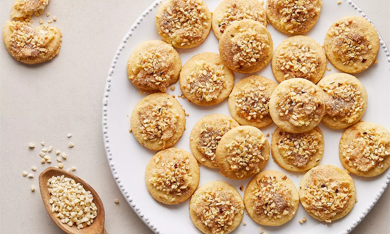 Yuvarlak, altın sarısı kurabiyeler beyaz tabakta düzenli sıralanmış, üzeri kıyılmış badem ve tarçın serpiştirilmiş, yanında tahta kaşıkta badem parçaları