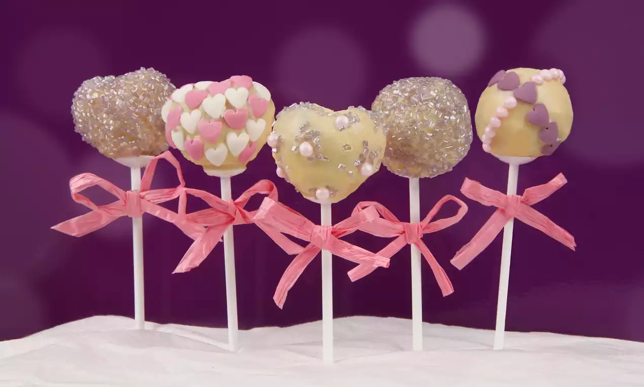 Šareni cake-popsi na bijelim štapićima, ukrašeni šljokicama, srcolikim konfetam i perlicama, s ružičastim vrpcama na ljubičastoj pozadini