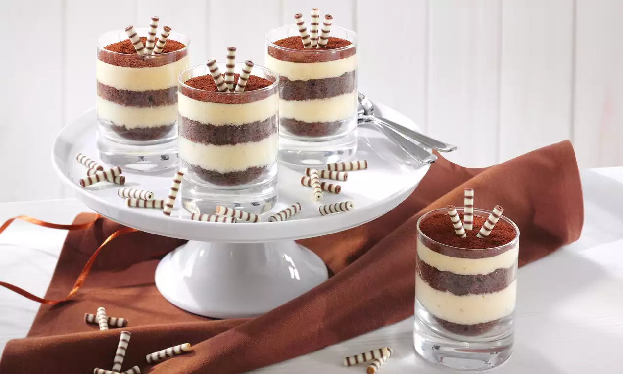 Schichtdessert im Glas mit abwechselnden Lagen aus heller Creme und dunklem Schokobiskuit, bestreut mit Kakaopulver und verziert mit gestreiften Schokoröllchen