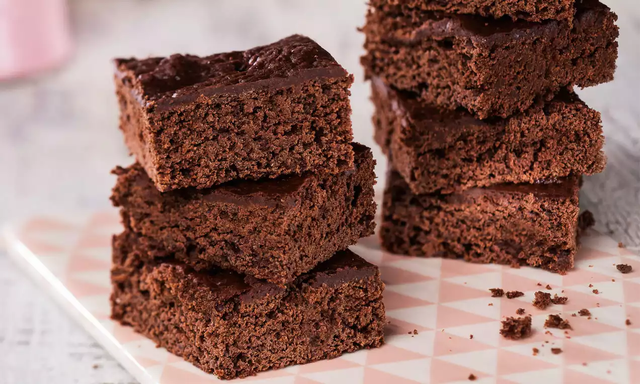 Dunkelbraune, quadratische Brownie-Stücke mit feuchter Textur und glatter Oberfläche, übereinander gestapelt auf einer rosa gemusterten Platte