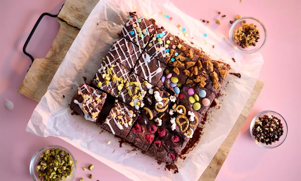 Donkerbruine brownies met decoraties van glazuur, kleurrijke sprinkles en toppings, gesneden en gerangschikt op bakpapier op een houten plank