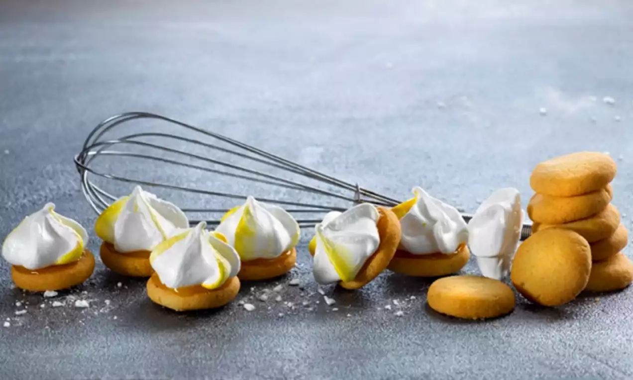 Keltaiset pikkuleivät, joiden päällä valkoiset marenkiviritykset ja sitruunankeltaista yksityiskohtaa, aseteltu vispilän viereen harmaalle pinnalle