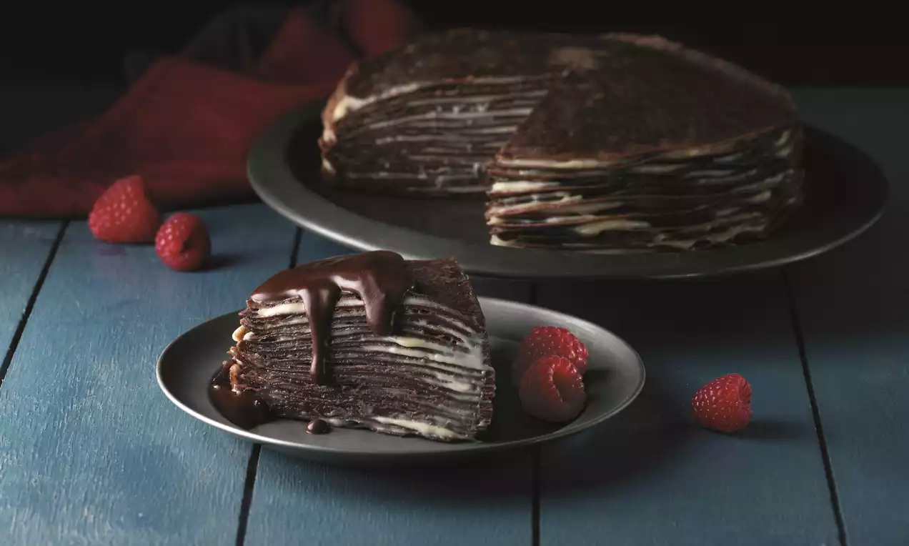 Layered pancake cake with chocolate-brown crepes and pale cream filling, slice topped with glossy chocolate sauce and raspberries on a dark plate
