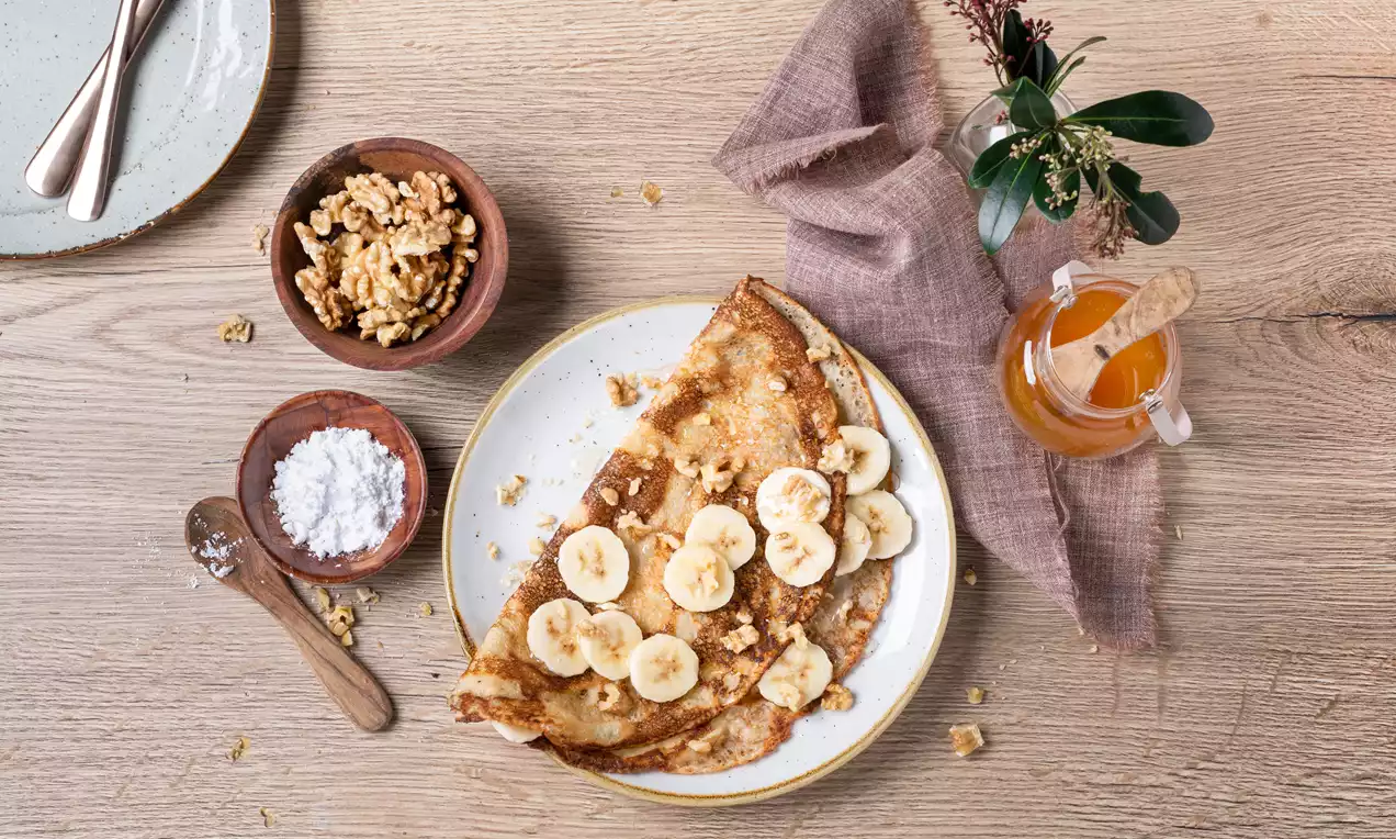 Crêpe dorée garnie de rondelles de banane jaune pâle, parsemée de noix concassées, présentée sur une assiette blanche bordée de jaune