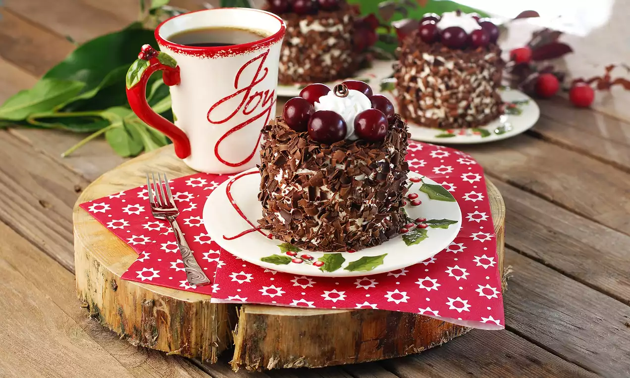 Küçük ahşap sunum tahtasında, kırmızı-yeşil motifli tabakta çikolata kaplı, vişne ve krema ile süslenmiş karaorman pastacıkları