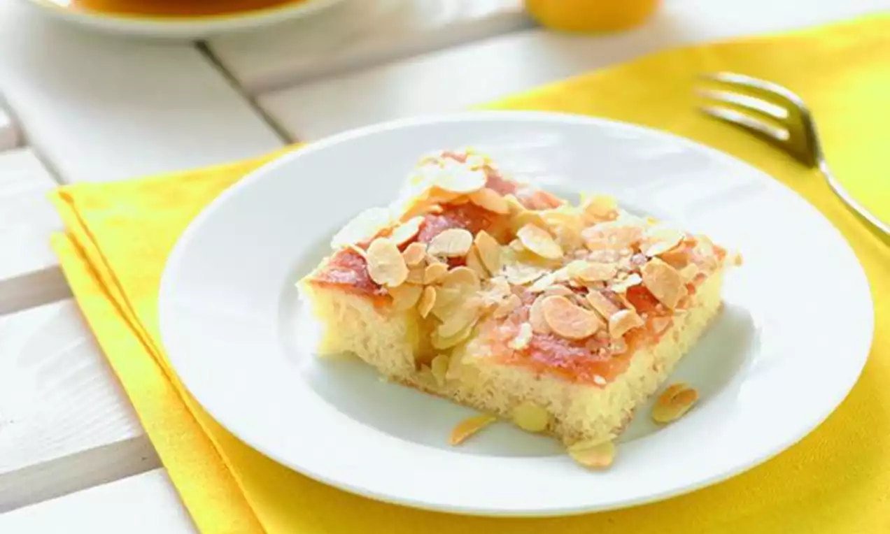 Firkantet smørkake med gyllen overflate og sprø mandelflak på en hvit tallerken på en gul duk med gaffel og kopp i bakgrunnen